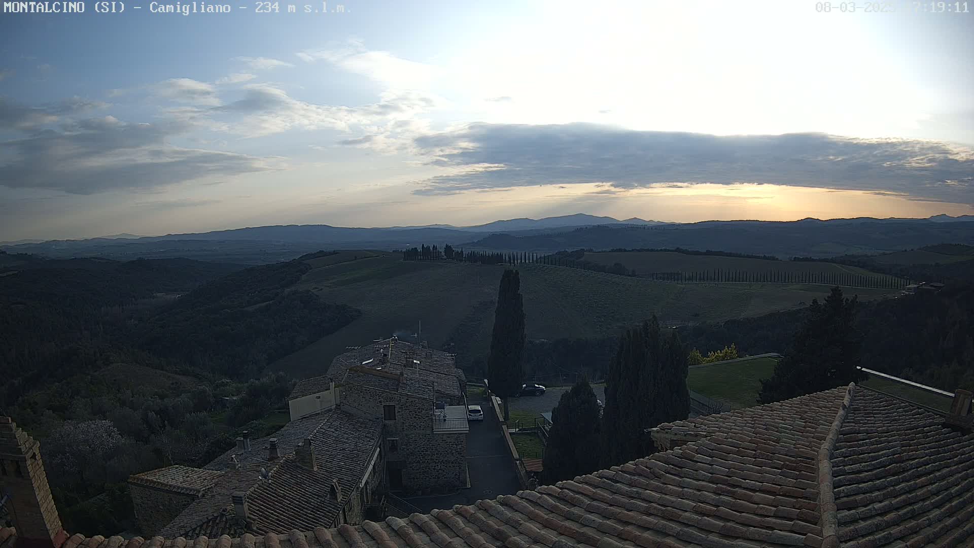 A sunlit, partly cloudy evening view of rolling hills and a village, seen from a rooftop with terracotta tiles.