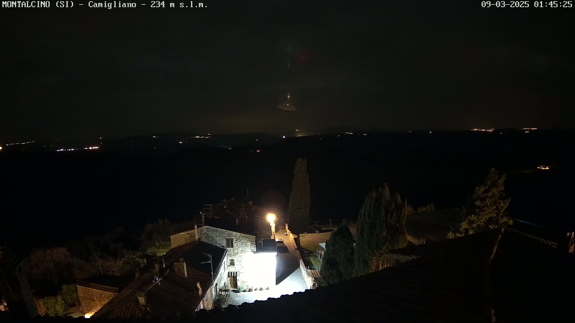 A nighttime, high-angle view shows a small village illuminated by lights, with a distant horizon showing faint lights and a dark, possibly cloudy sky.