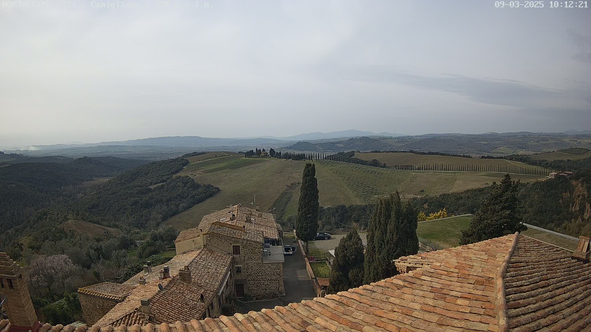 A view from a terracotta roof shows a stone village nestled in rolling hills under a hazy sky.