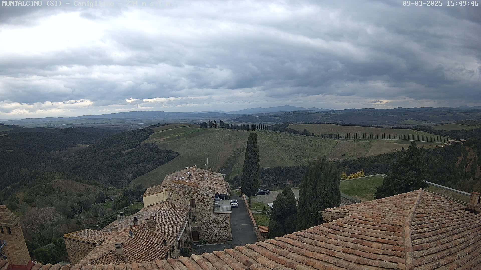 A view from a rooftop shows rolling hills under a cloudy sky, with stone buildings and cypress trees in the foreground.