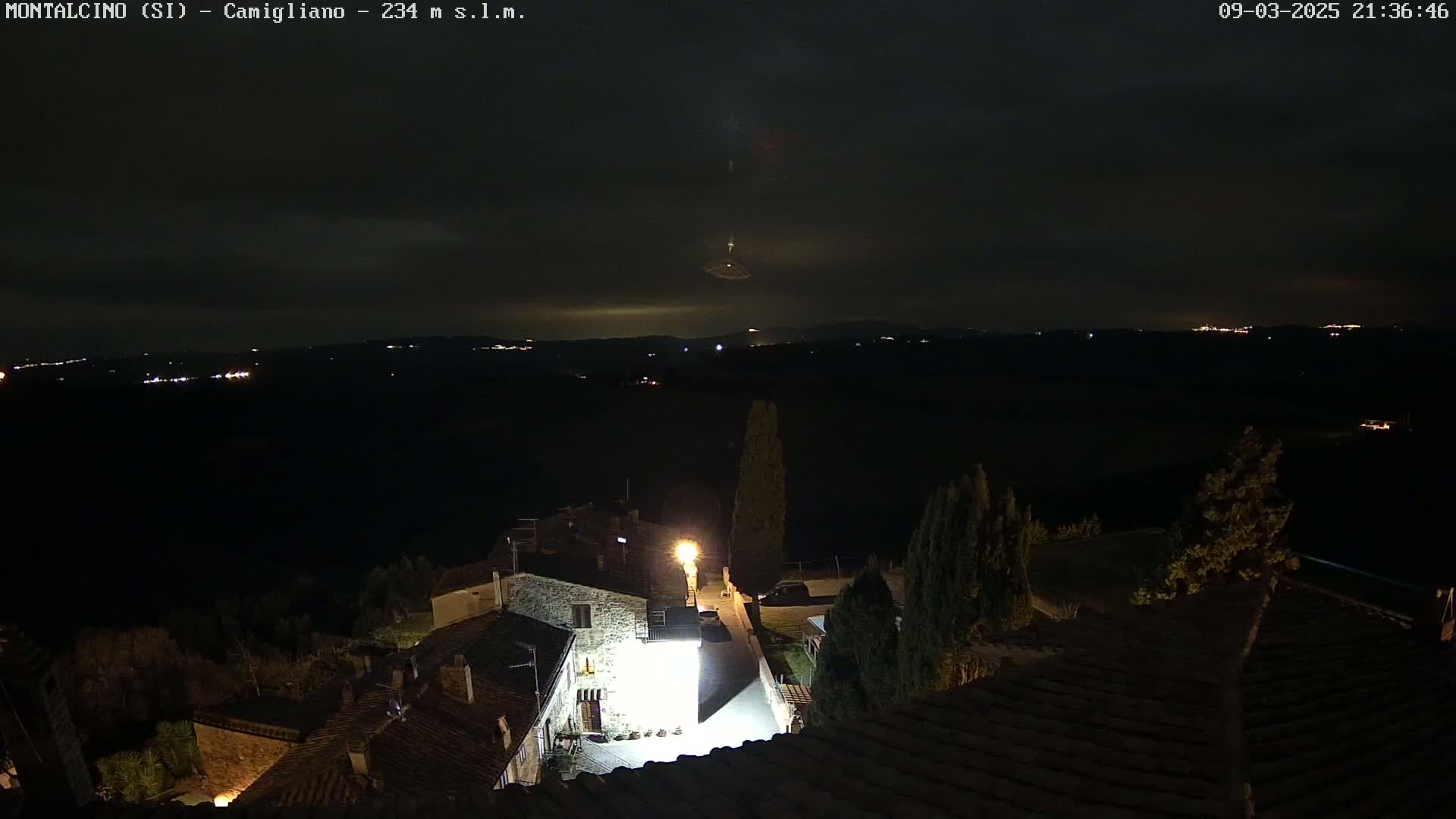 A nighttime view of a small village nestled in a hilly landscape under a dark, cloudy sky with a faint light display in the distance.