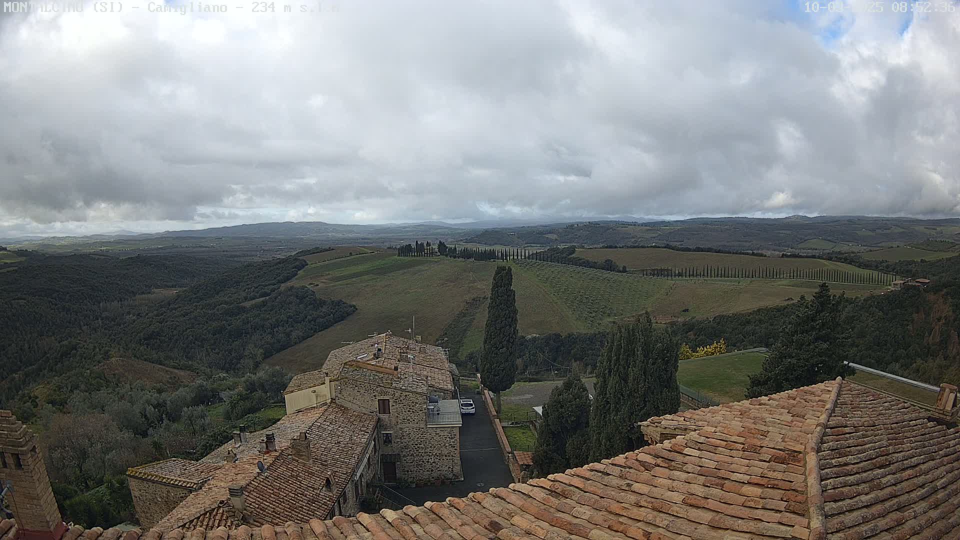A view from a rooftop shows rolling hills under a cloudy sky, with stone buildings and cypress trees in the foreground.