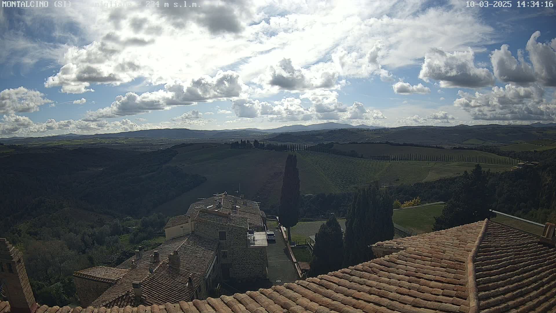 A partly cloudy sky overlooks a hilly landscape of rolling green fields, cypress trees, and terracotta-roofed buildings.
