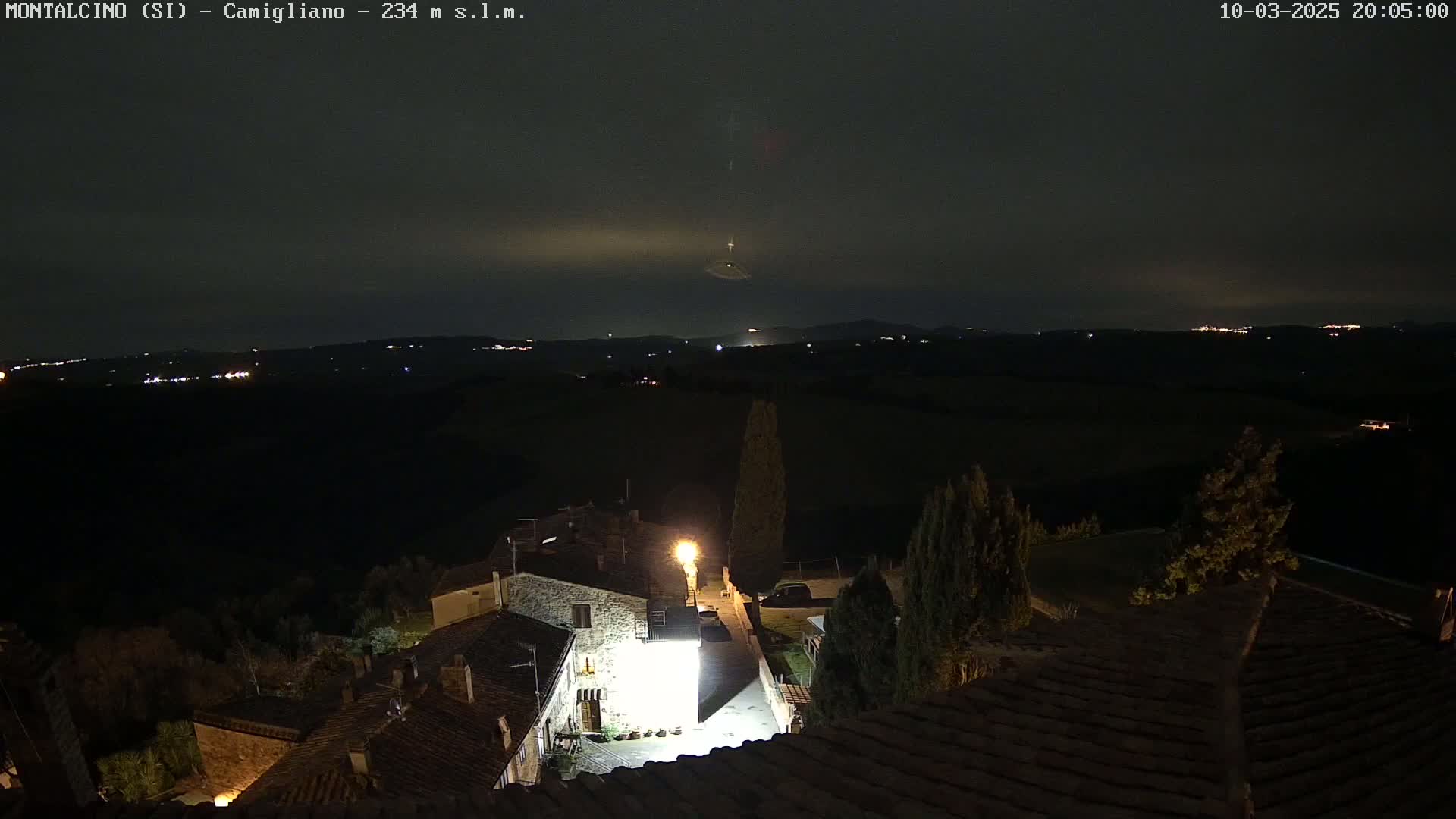 A nighttime view of a small village with stone buildings, illuminated by streetlights, overlooking a dark, hilly landscape under a mostly clear sky.