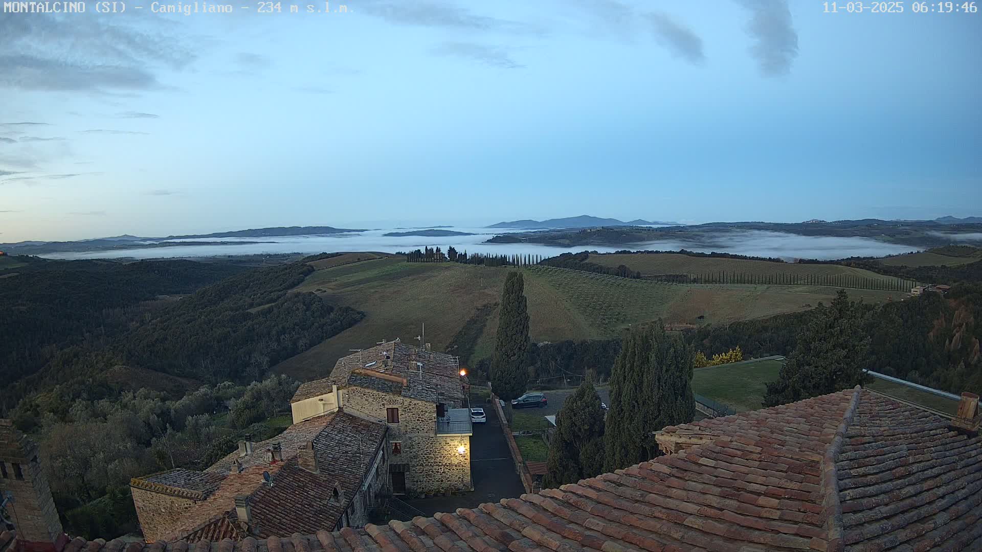 A low-lying fog covers a valley below a hilltop village at dawn, with stone buildings and terracotta roofs visible in the foreground.
