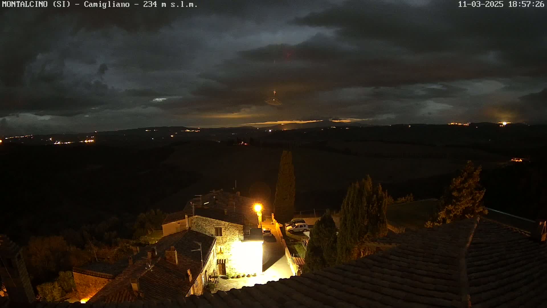 A nighttime view of a village nestled in a hilly landscape under a dark, cloudy sky, with a few lights visible in the distance.