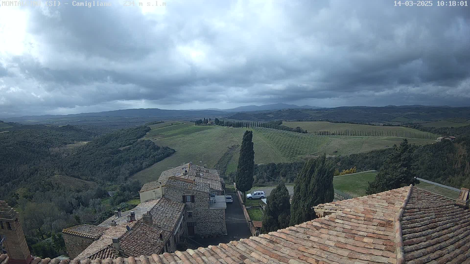 Under a cloudy sky, a view from a rooftop shows stone buildings and a rolling landscape of green hills and cypress trees.