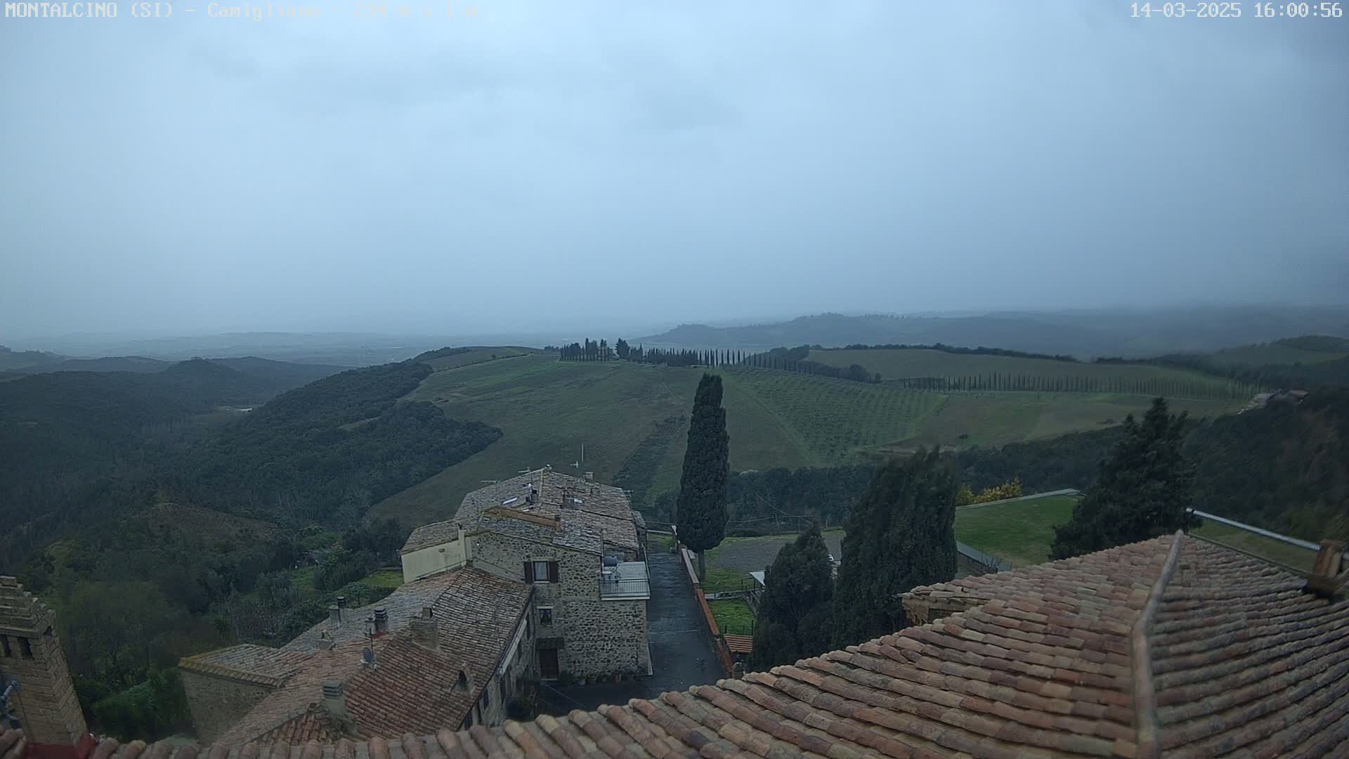 Overcast skies reveal a view of rolling green hills and vineyards, a village with terracotta rooftops nestled among the landscape.