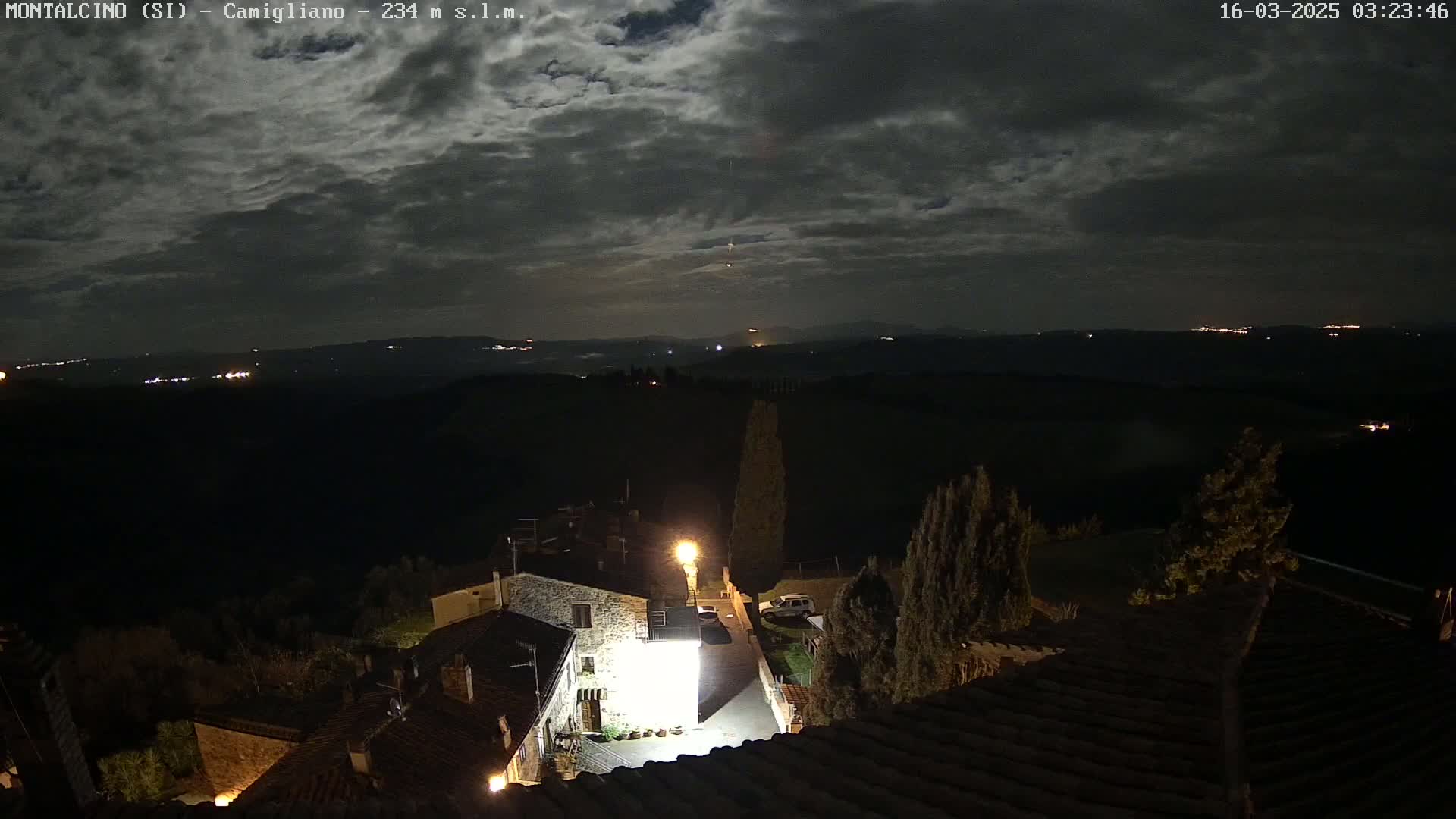 A nighttime view of a small village nestled in a hilly landscape under a partly cloudy moonlit sky.
