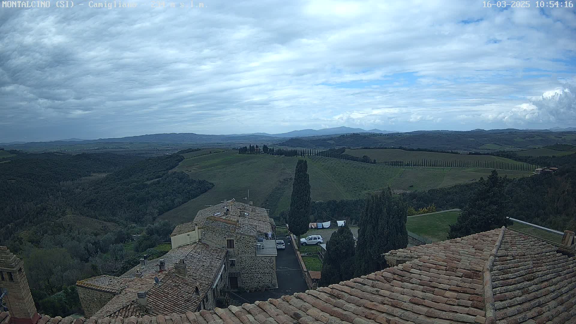 A view from a rooftop shows a partly cloudy sky over rolling green hills and distant mountains, with stone buildings and terracotta roofs in the foreground.