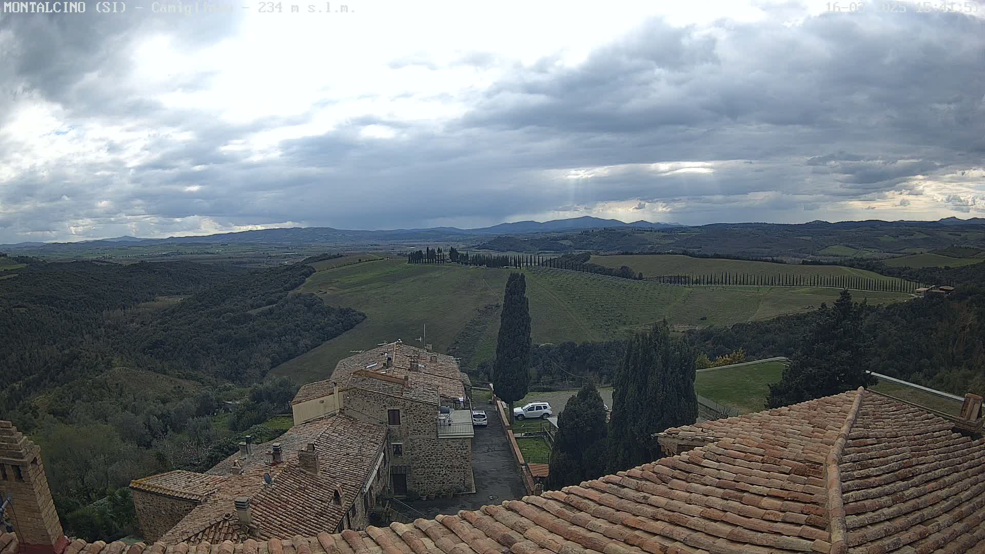 Overlooking rolling hills and a village under a cloudy sky, terracotta rooftops are in the foreground.