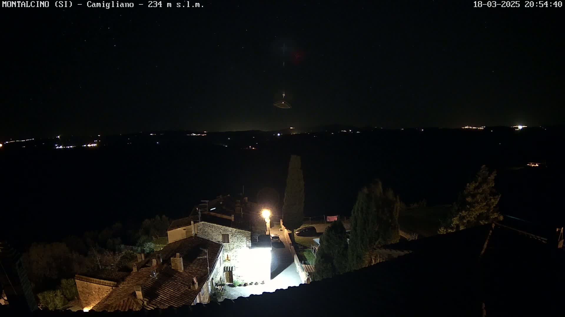 A nighttime, clear-sky view of a small village illuminated by streetlights, with distant lights visible on a hillside in the background.