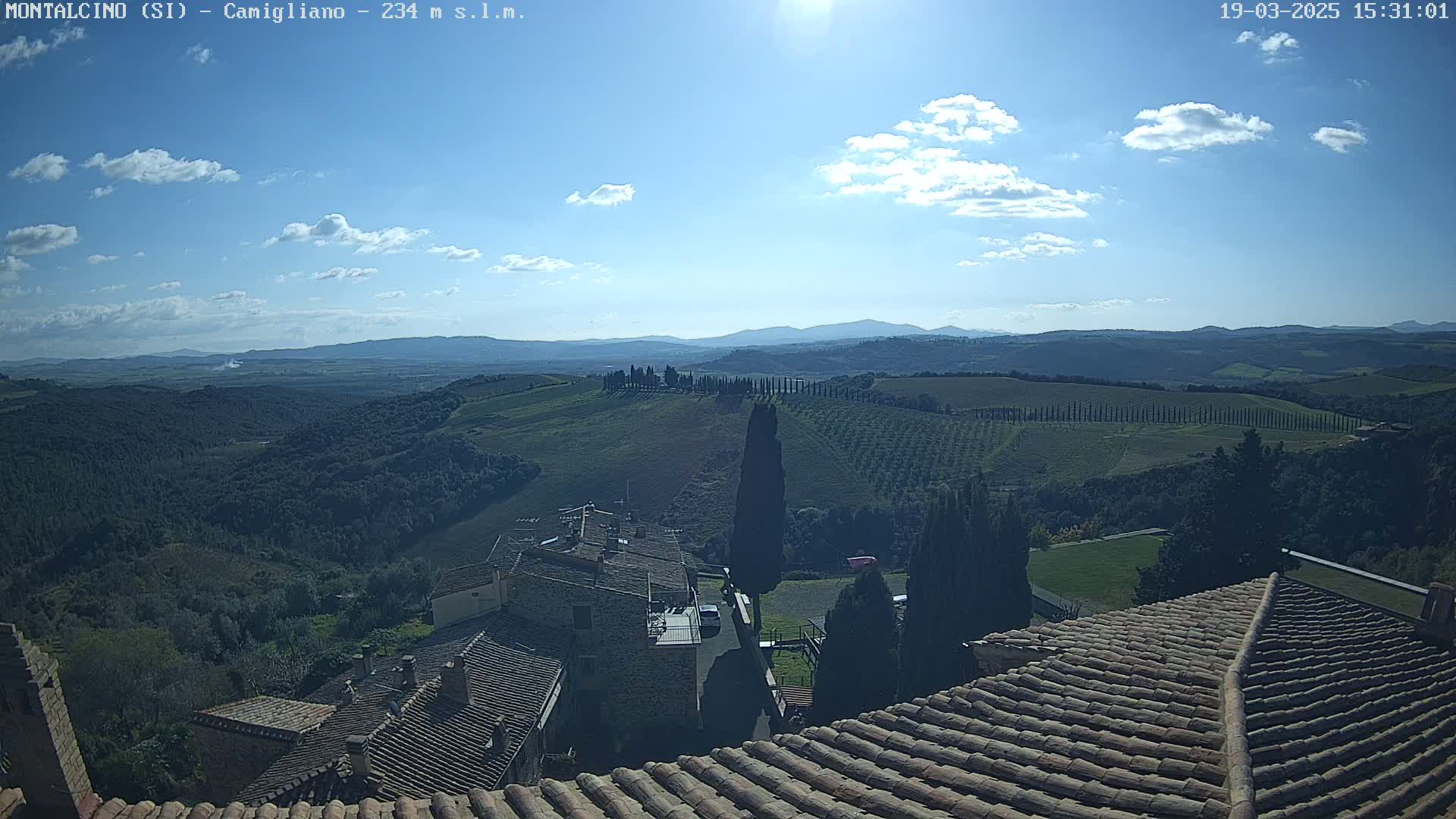 A sunny day reveals a sprawling Tuscan landscape of rolling hills, vineyards, and cypress trees, viewed from a rooftop overlooking a village of stone buildings.