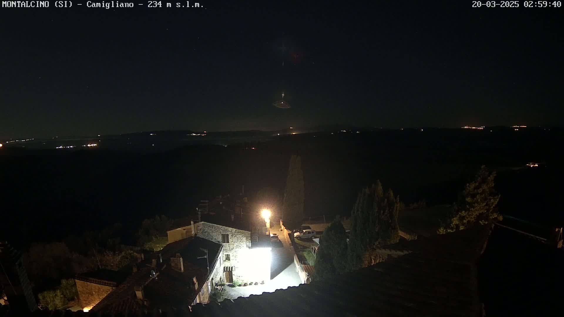 A nighttime, high-angle view of a small village illuminated by streetlights, with a dark, starlit sky and distant lights visible on the horizon.