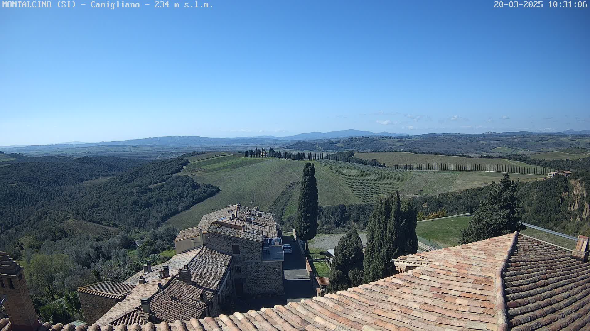 A sunny day reveals a picturesque Tuscan landscape, showcasing rolling green hills dotted with cypress trees, a village with terracotta roofs, and distant mountains under a clear blue sky.