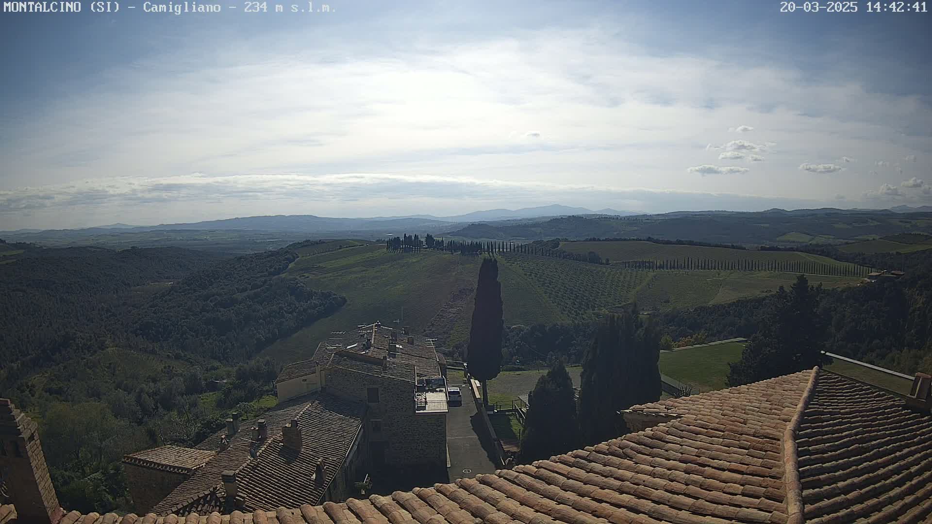 A view from a terracotta roof shows a partly cloudy sky over a hilly landscape of olive groves and stone buildings under a partly cloudy sky.