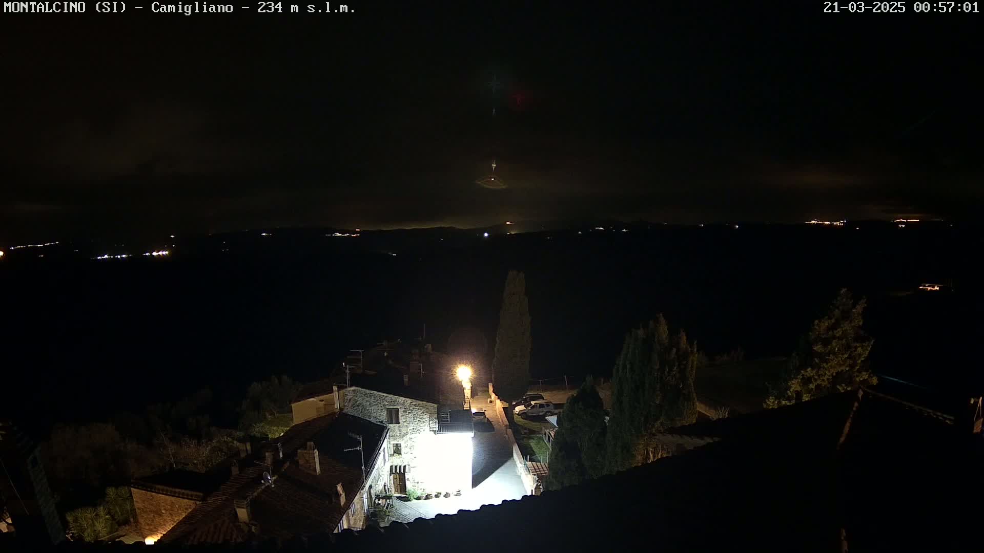 A nighttime, high-angle view shows a small village with dimly lit buildings under a mostly clear, dark sky, with distant lights visible on a horizon over a valley.