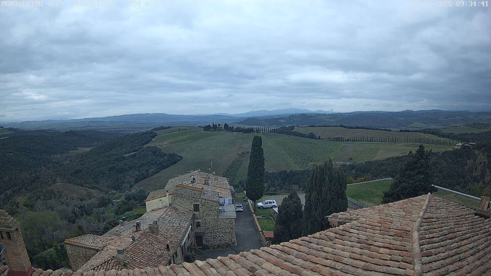 Stone buildings with terracotta roofs overlook a sprawling landscape of rolling hills, vineyards, and cypress trees under a cloudy sky.