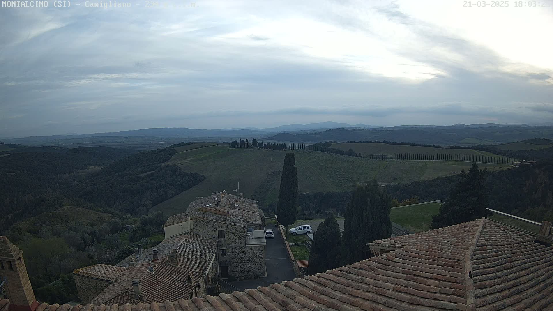 A view from a rooftop shows a cloudy sky over rolling hills,  with a stone village nestled in the foreground.