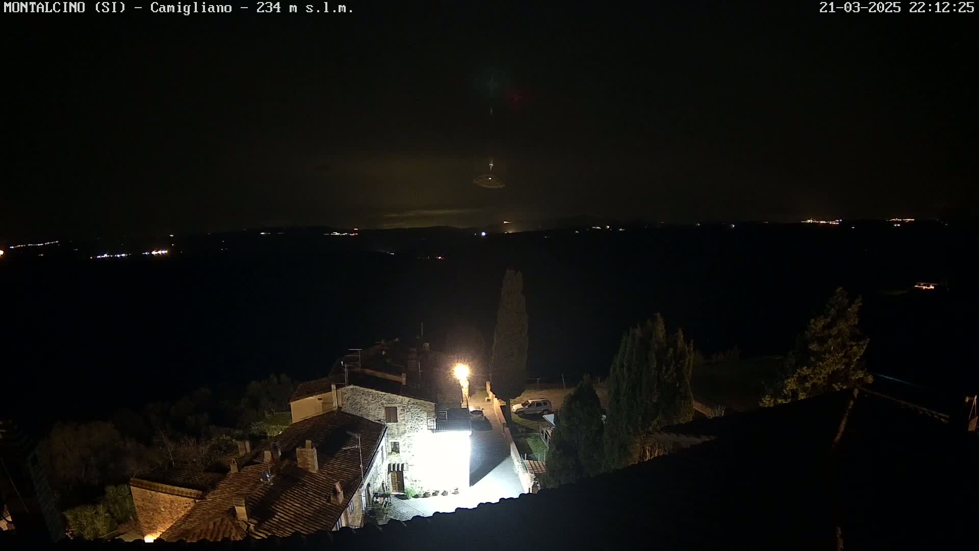 A nighttime view of a small village illuminated by lights, with a distant horizon showing faint lights and a possible meteor shower in a dark, clear sky.