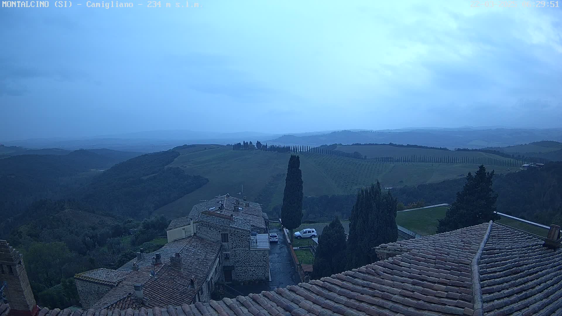 Overcast, hilly farmland and a village with terracotta roofs are viewed from above.