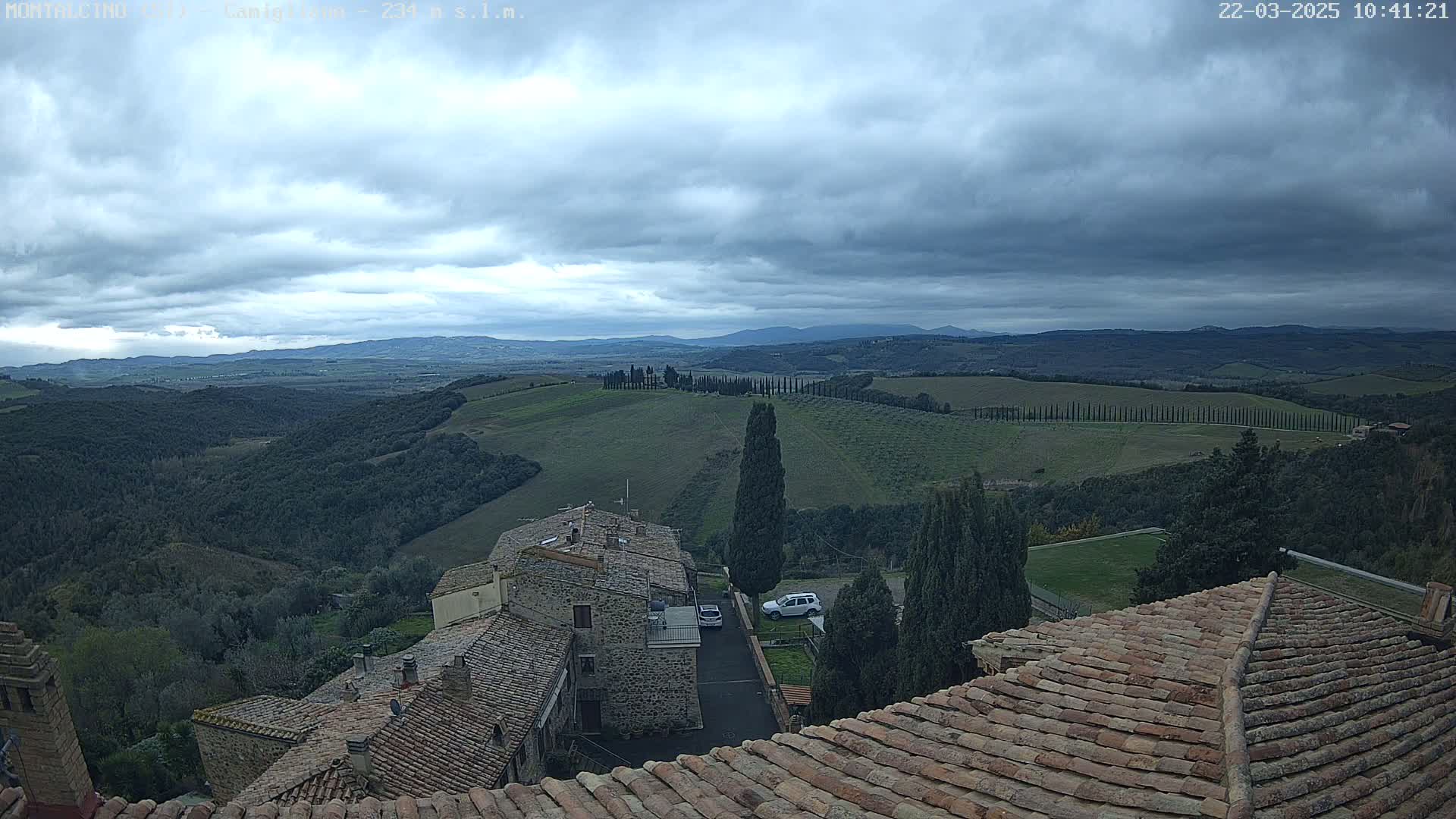 A view from a rooftop shows a cloudy day over rolling green hills dotted with cypress trees and stone buildings, with distant mountains visible on the horizon.
