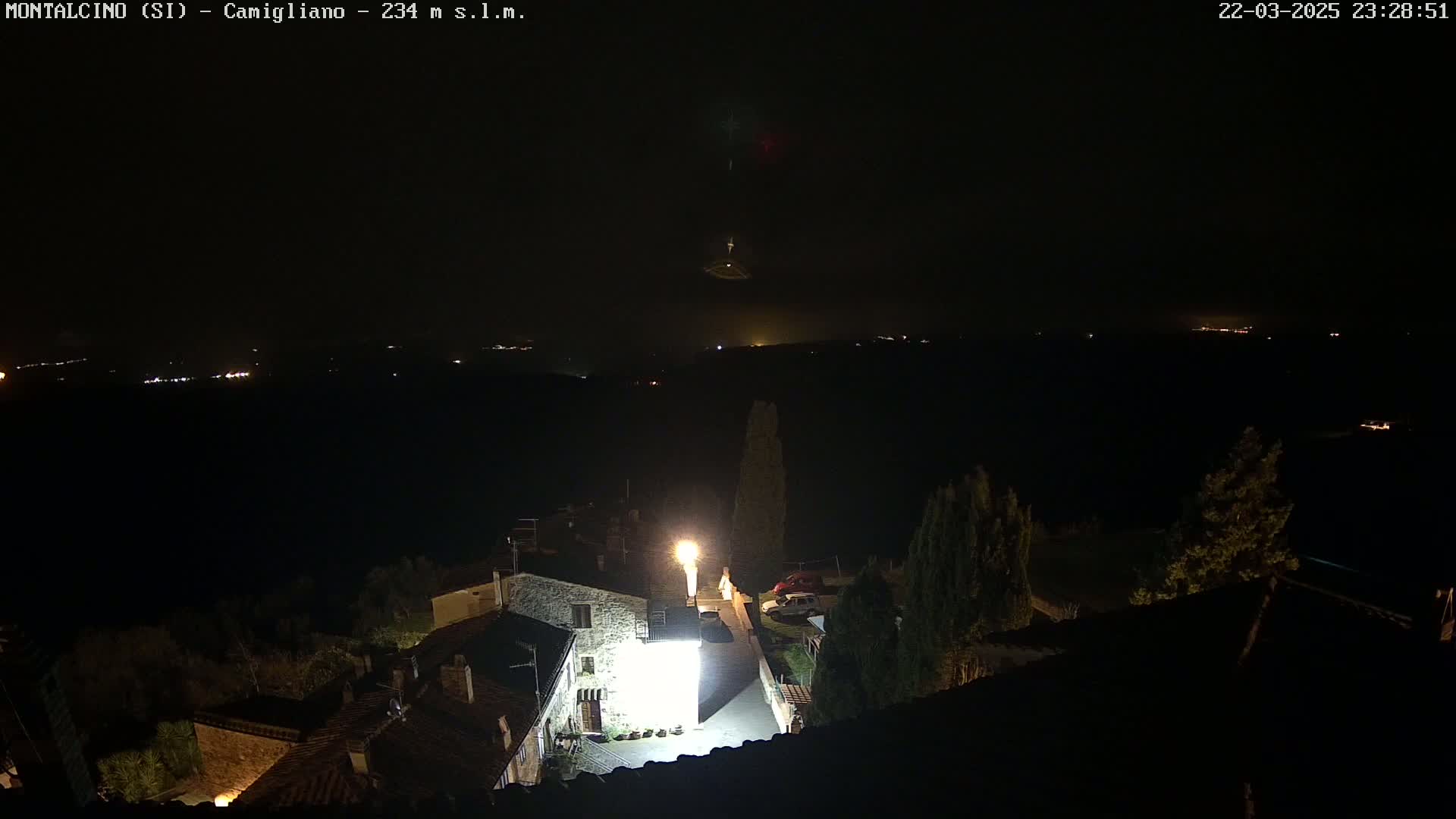 A nighttime view of a small village illuminated by streetlights, with a dark, distant landscape and a clear sky visible in the background.