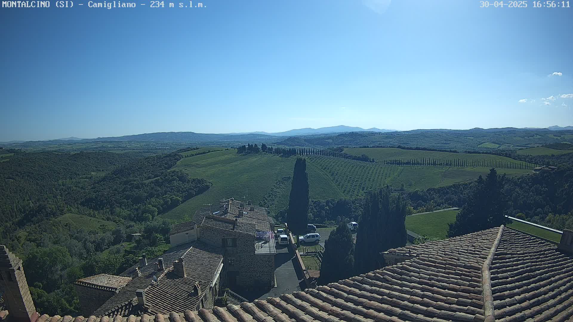 A clear, sunny view from a rooftop shows a picturesque Tuscan landscape of rolling green hills, cypress trees, and a stone village, with distant mountains visible on the horizon.