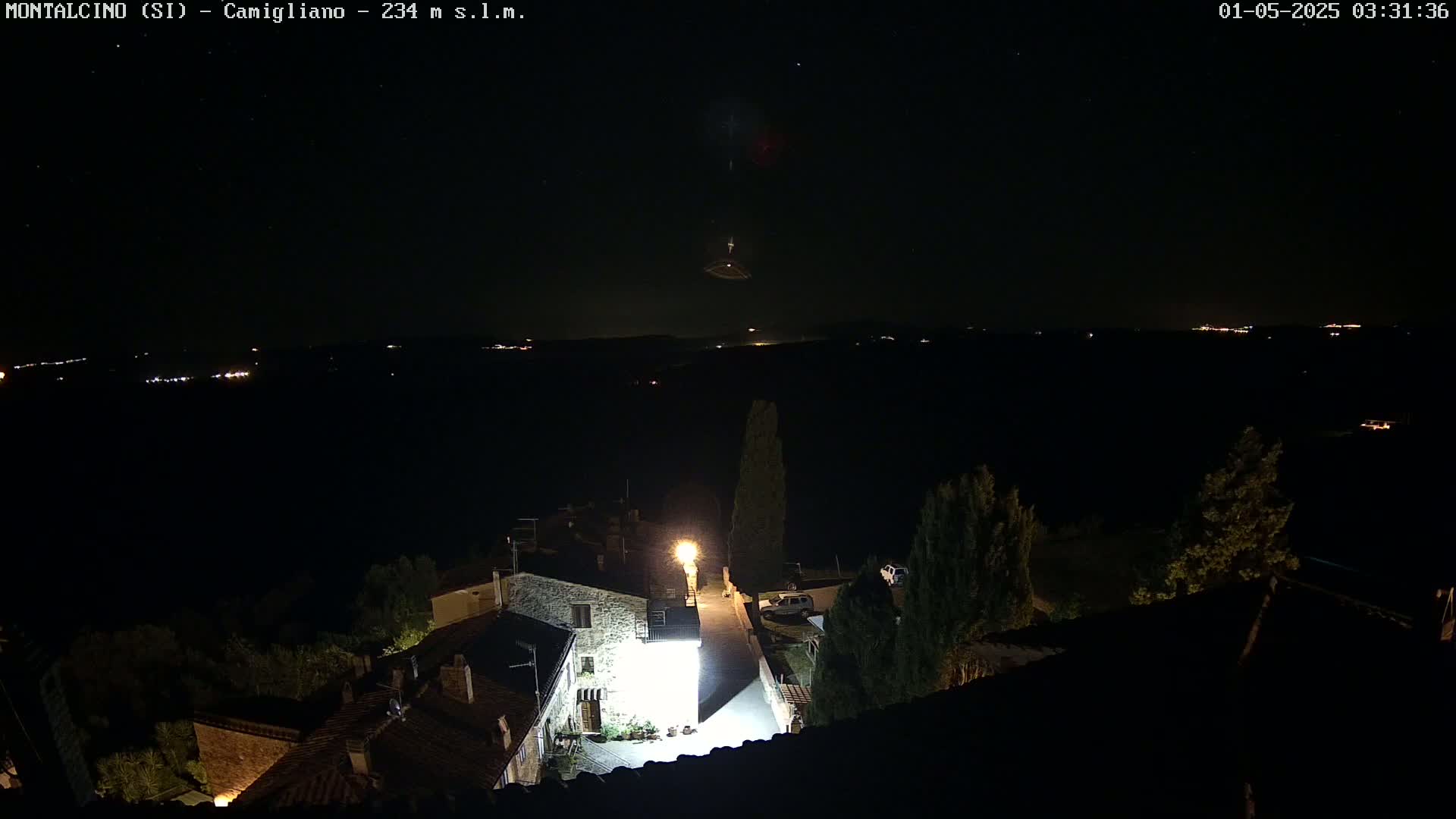 A nighttime, high-angle view of a small village illuminated by streetlights, with a dark, starlit sky and distant lights on the horizon.