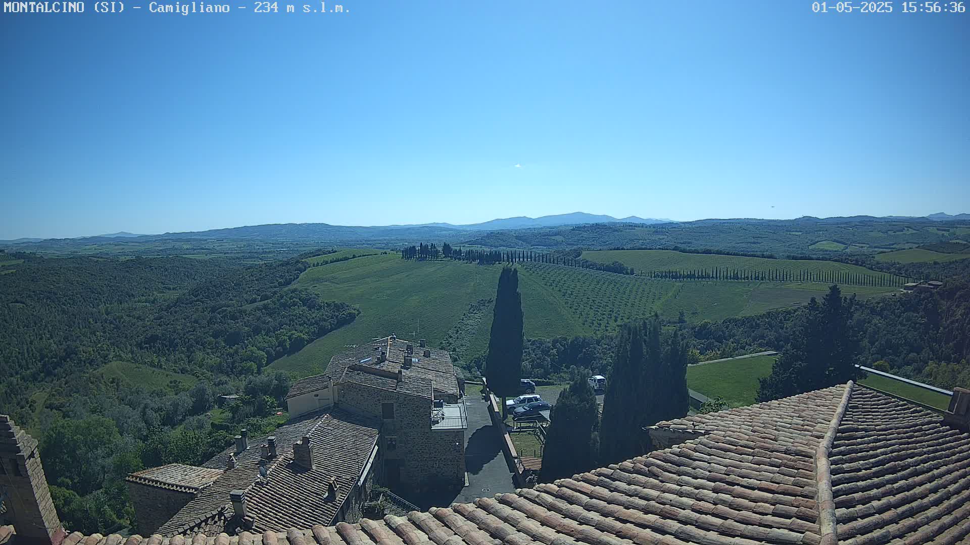 A sunlit view from a terracotta roof shows a stone village nestled in rolling green hills under a clear blue sky.