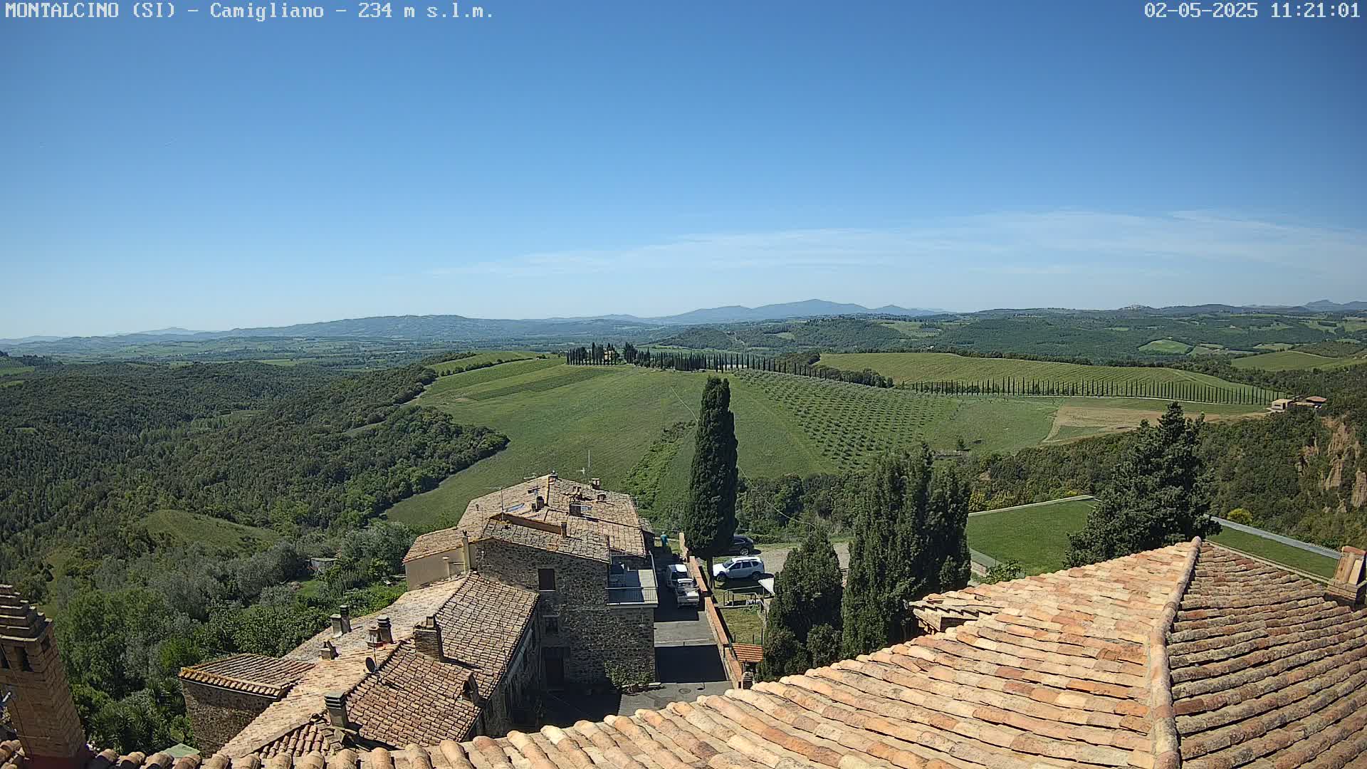 A sunny view from a rooftop shows terracotta-tiled roofs of buildings overlooking a rolling green landscape with rows of trees and distant mountains under a clear blue sky.