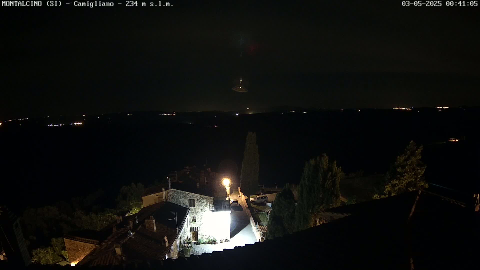 A nighttime, high-angle view shows a small village illuminated by streetlights, with a dark, distant landscape and a faint light display in the sky.