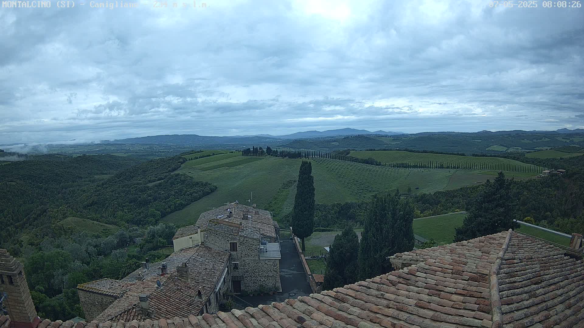Overcast skies reveal a panoramic view of rolling green hills, dotted with cypress trees and topped with terracotta rooftops of buildings.