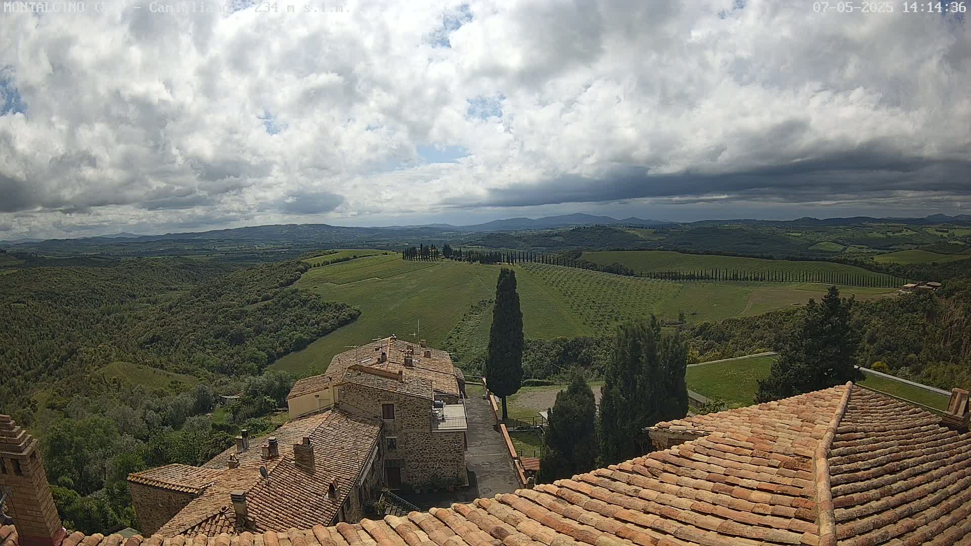 Stone buildings with terracotta roofs overlook a partly cloudy landscape of rolling green hills and distant mountains.