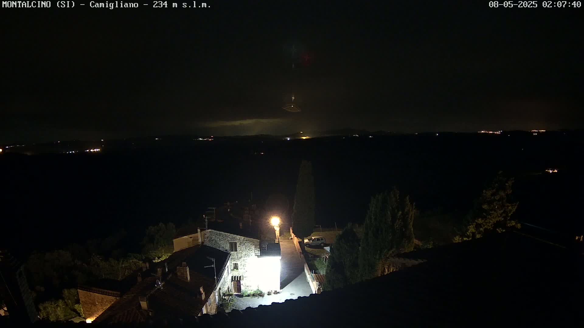 A nighttime view of a small village illuminated by a single bright light, overlooking a dark landscape under a partly cloudy sky with faint lightning in the distance.