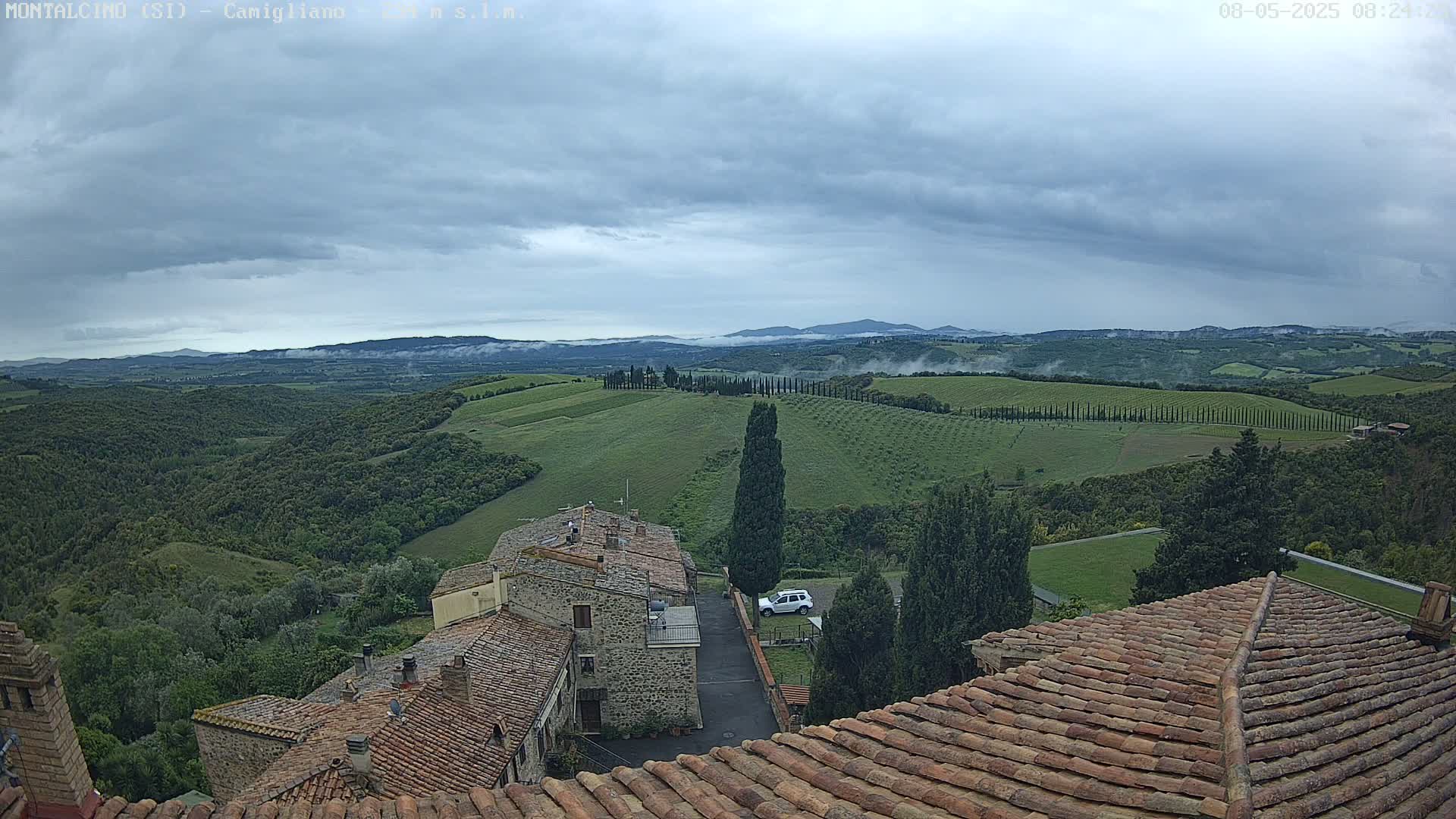 Overcast skies loom over a hilly landscape of green fields, cypress trees, and stone-roofed buildings, with a sliver of fog in the distance.