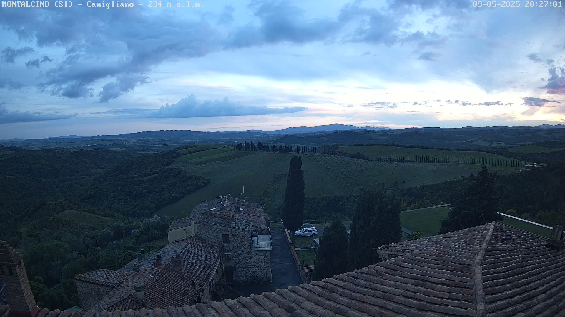 Stone rooftops of buildings overlook a vast, rolling landscape of green hills and olive groves under a partly cloudy, twilight sky.