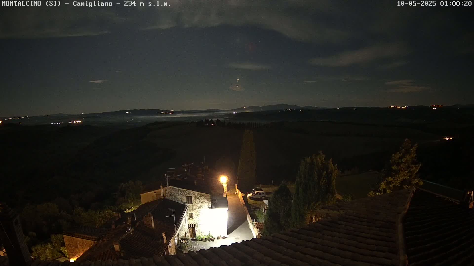 A nighttime view of a small village nestled in a hilly landscape under a partly cloudy sky, with low-lying fog in the distance.