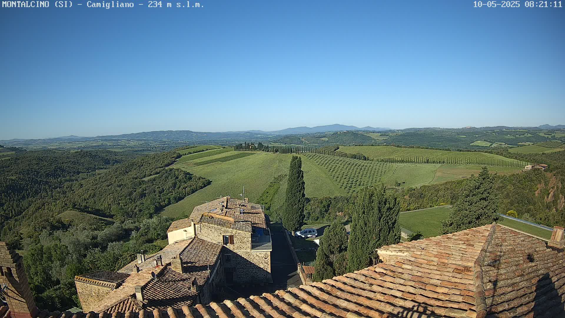 A clear, sunny day reveals a panoramic view of rolling green hills, dotted with trees and farmlands, overlooking terracotta rooftops of buildings.