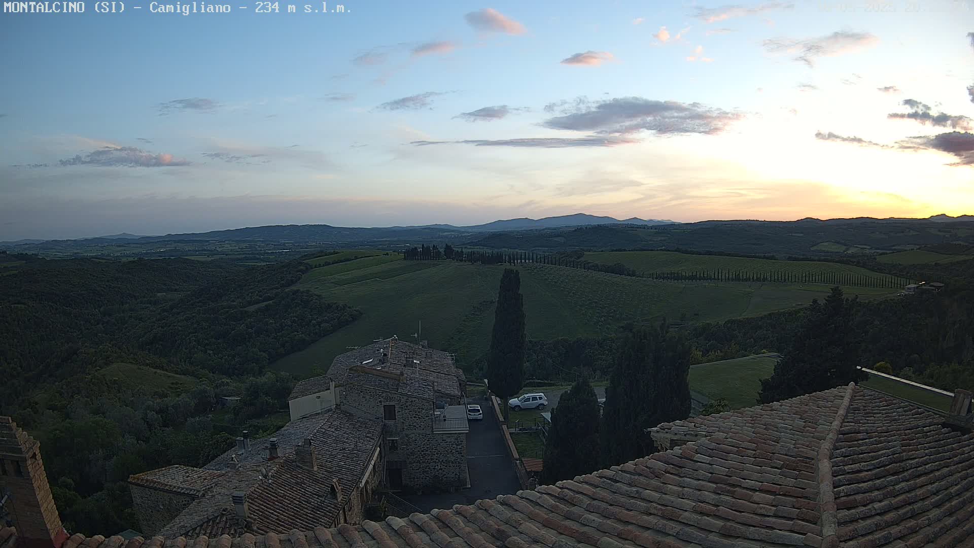 Under a partly cloudy sunset sky, a view from a rooftop shows stone buildings and rolling green hills extending to distant mountains.