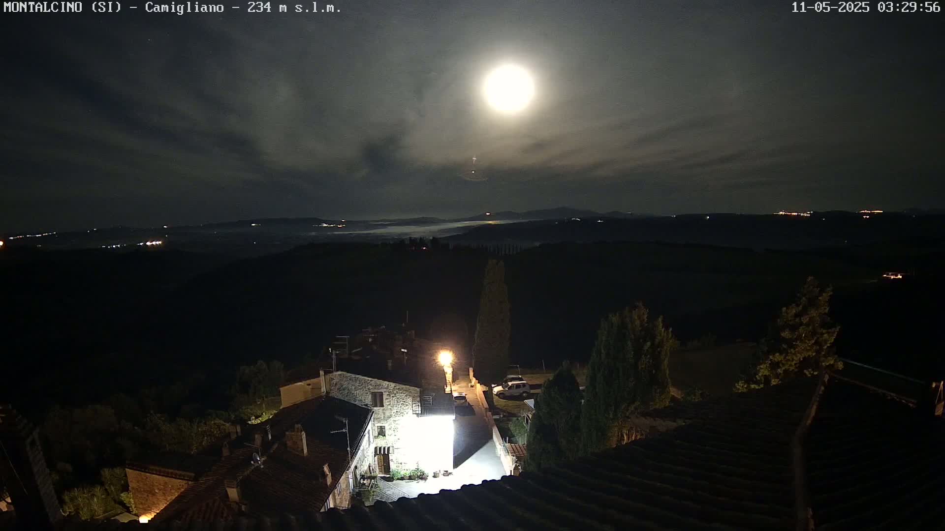 A brightly lit moon shines over a dark, hilly landscape at night, illuminating a small village with stone buildings.