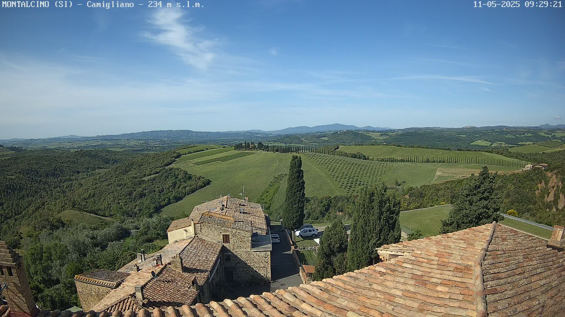 A view from a terracotta roof shows a sunny, mostly clear day over rolling green hills dotted with trees and buildings, with distant mountains in the background.