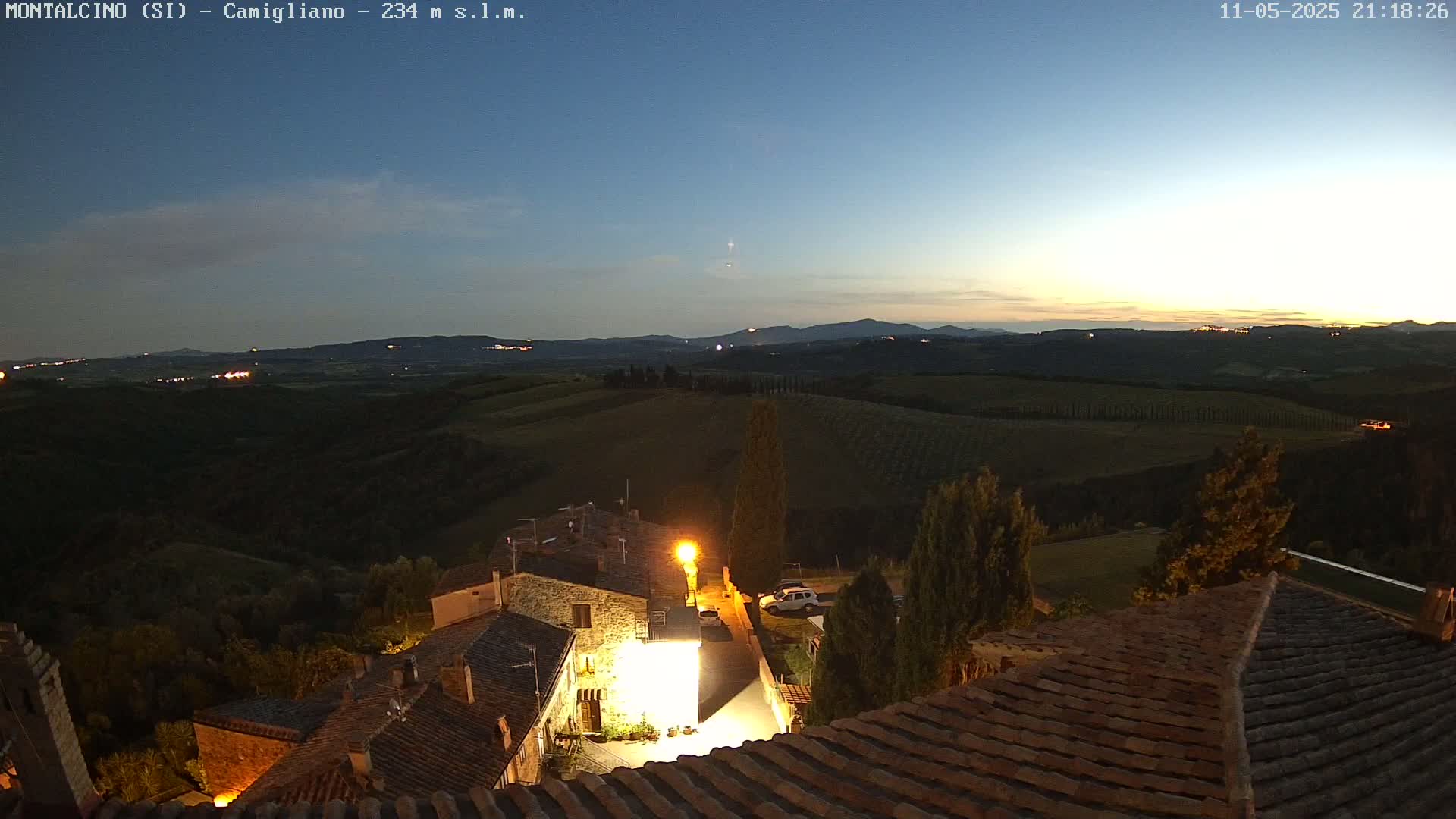A nighttime view of a small Italian village nestled in rolling hills under a twilight sky.