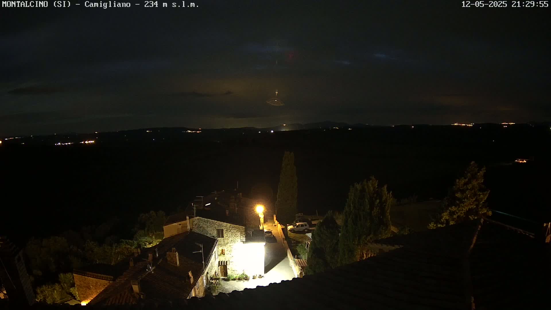 A nighttime view of a small village nestled in a dark landscape under a mostly clear sky, with a few distant lights visible on the horizon.
