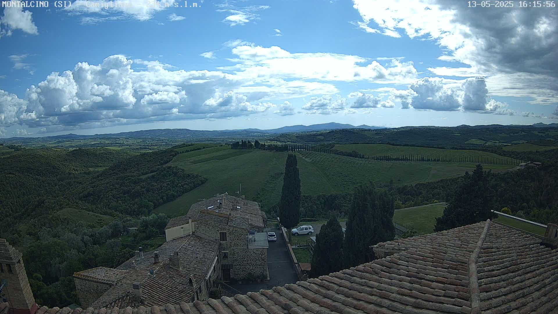 A partly cloudy sky overlooks rolling green hills and the terracotta roofs of a village.