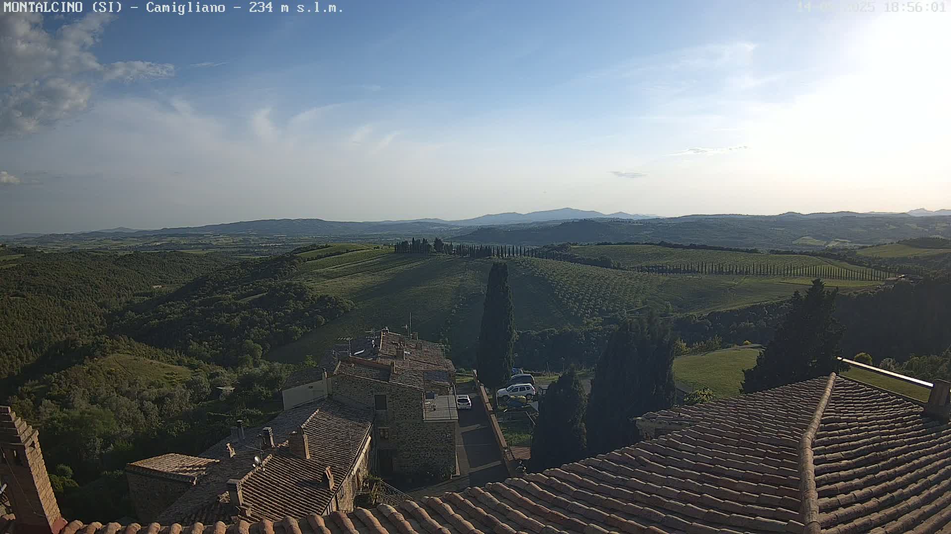 A view from a tiled rooftop shows a sunlit Tuscan landscape of rolling green hills, dotted with trees and buildings, under a mostly clear sky.