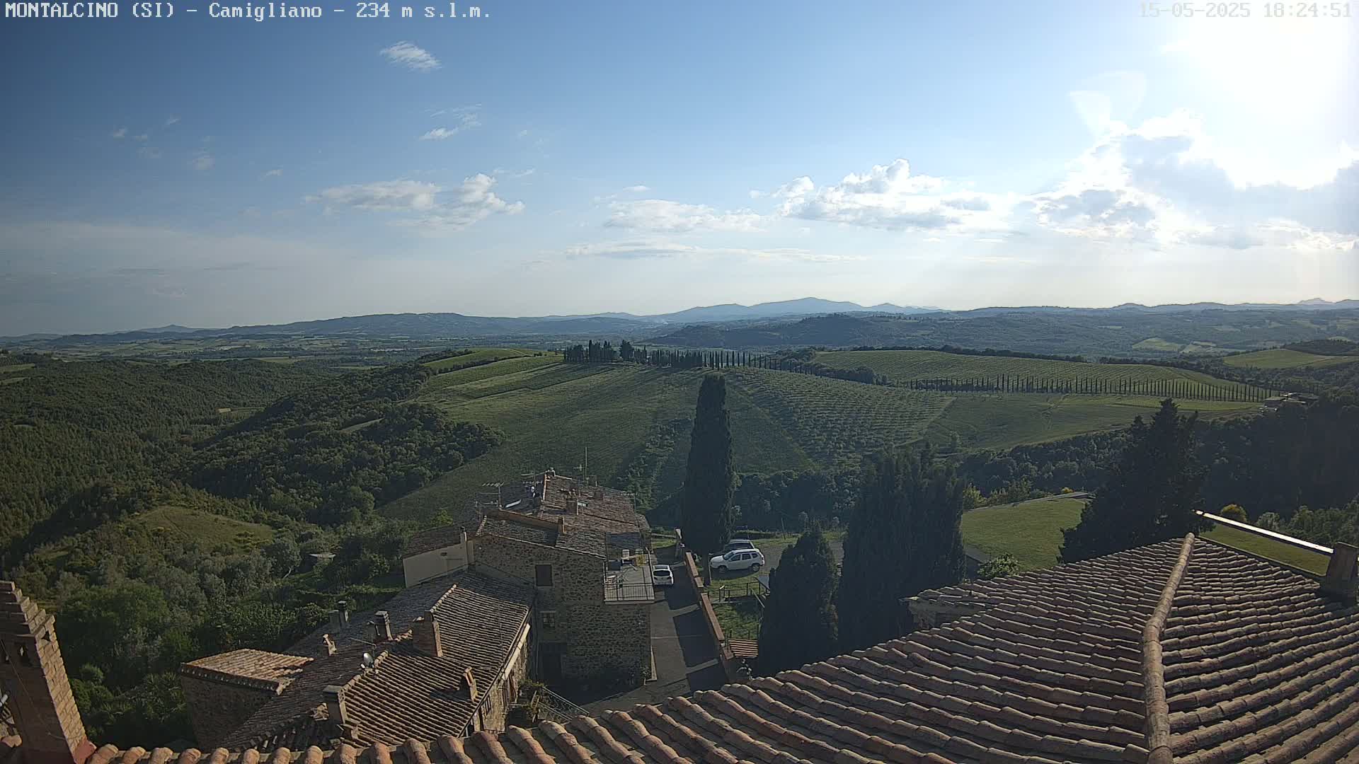 A partly cloudy daytime view of rolling green hills and a village with terracotta rooftops, seen from above.