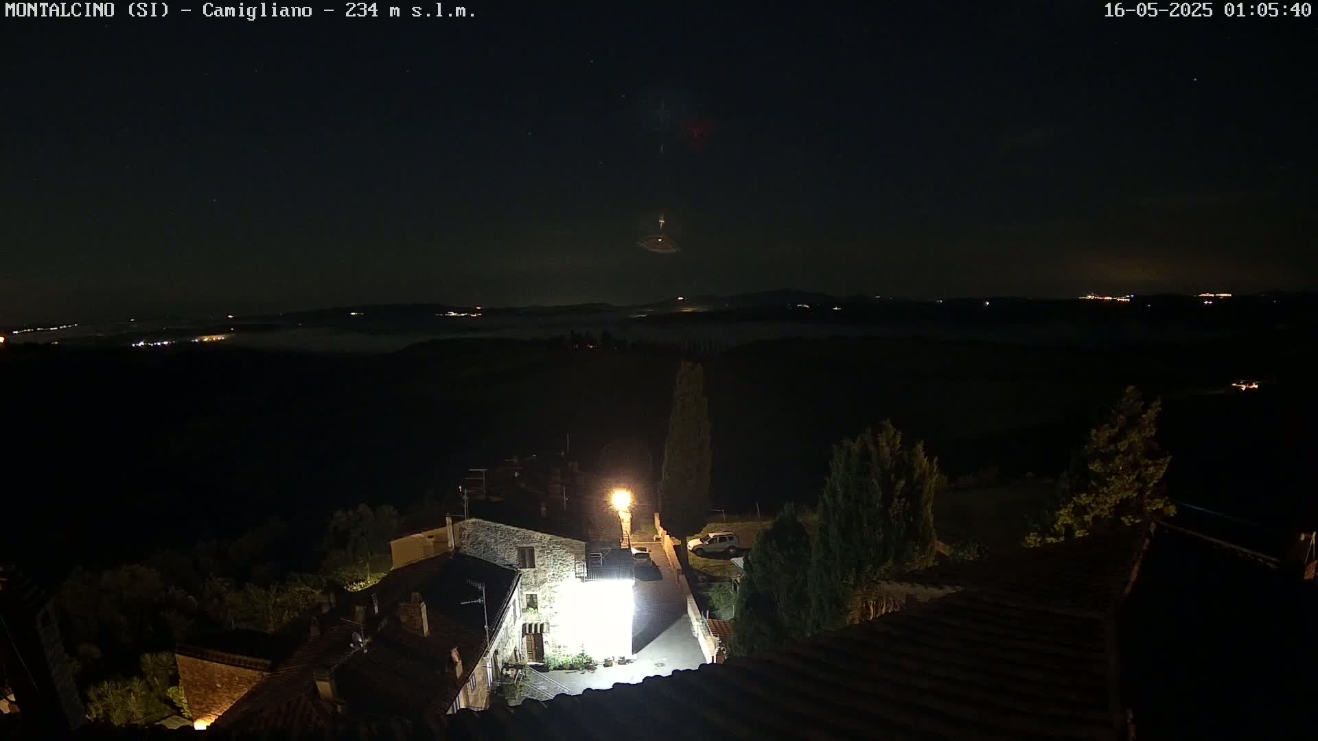 A nighttime view of a small village nestled in a valley, illuminated by streetlights, overlooks a low-lying layer of fog with distant lights visible under a dark, starry sky.