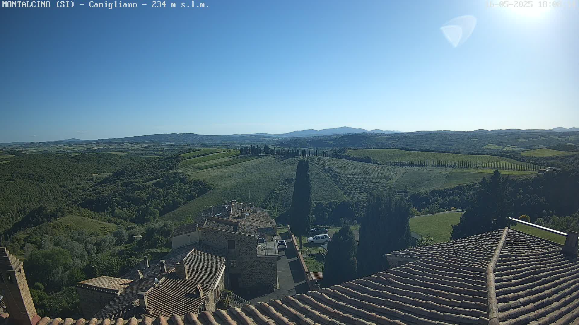 Under a clear blue sky, a view from a rooftop shows a sprawling landscape of rolling green hills, vineyards, and a small village, with distant mountains in the background.
