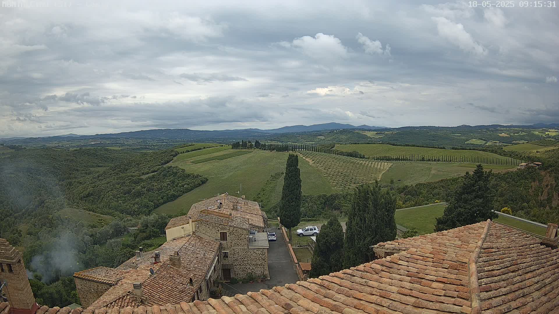 A view from a rooftop shows rolling hills of farmland under a cloudy sky, with several stone buildings and a few cars visible in the foreground.